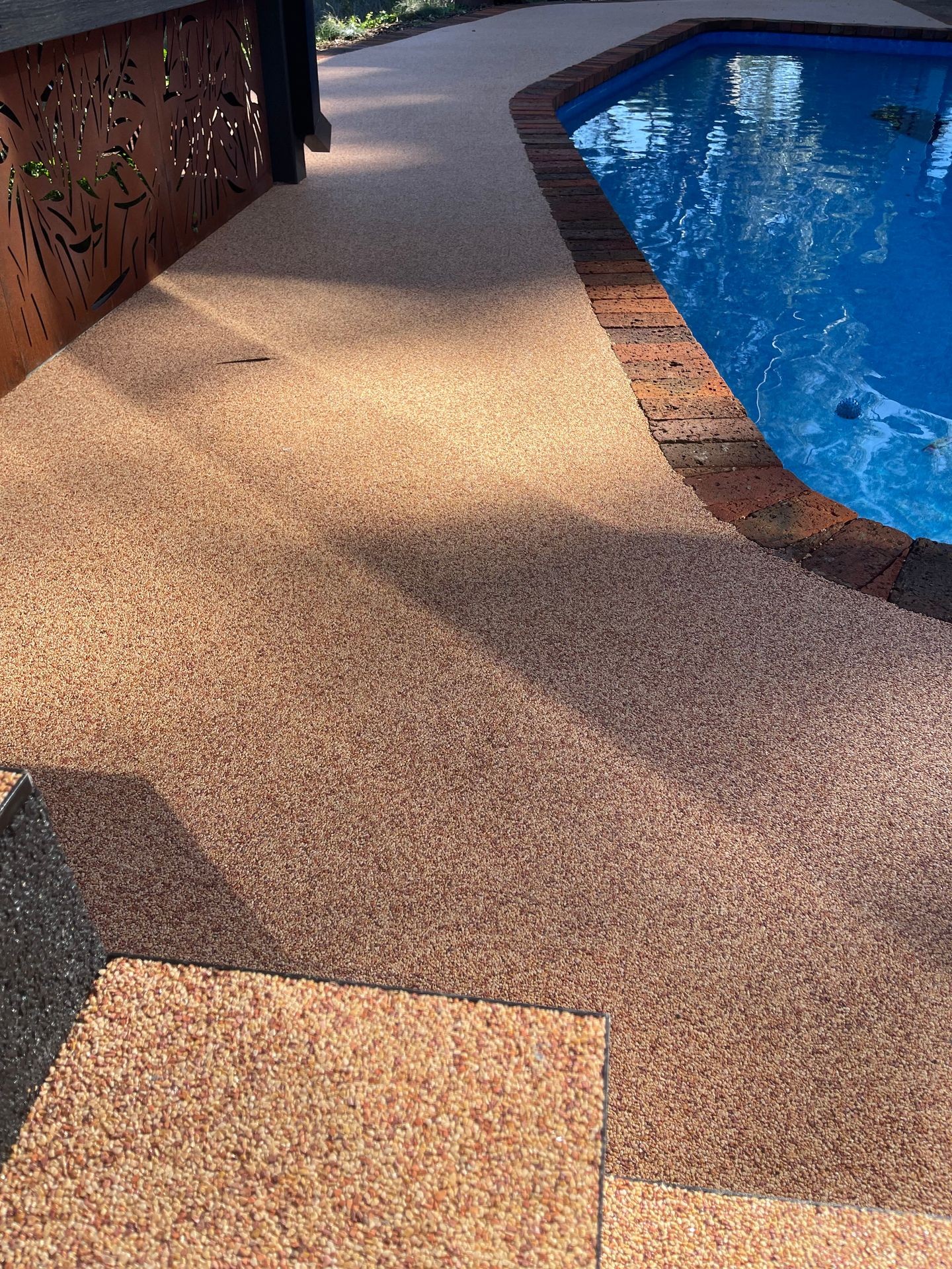 Outdoor swimming pool with textured beige pathway and brick border in sunlight.
